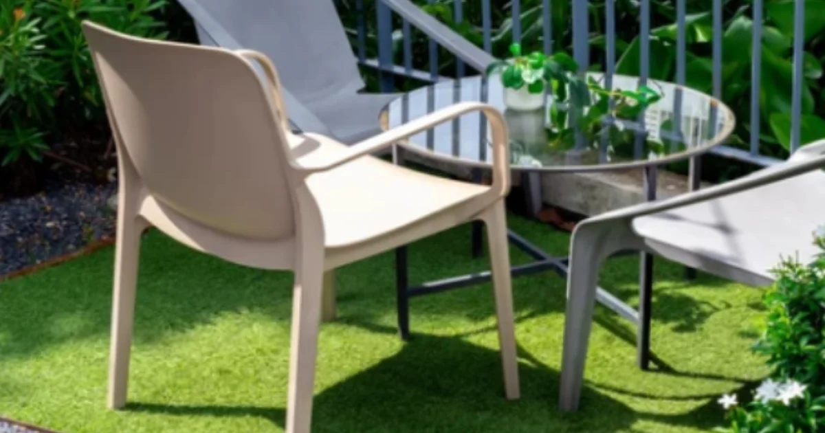 Outdoor patio seating area with beige and gray plastic chairs around a glass table set on artificial turf.